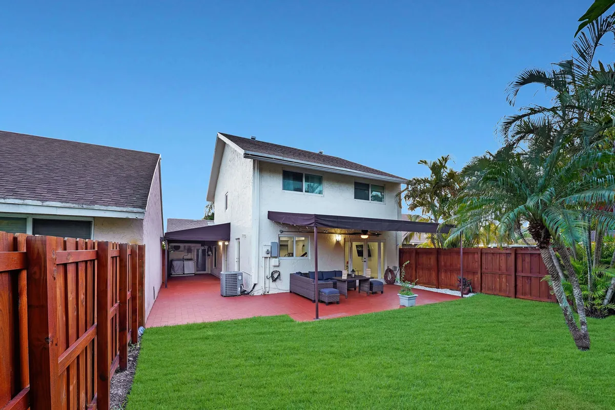 This exterior image shows a spacious backyard with a patio seating area, green lawn, decorative plants, and a fenced perimeter. The mood is welcoming and relaxing, ideal for outdoor dining or family gatherings. The lush greenery and open space underscore a sense of tranquility and work/life balance.