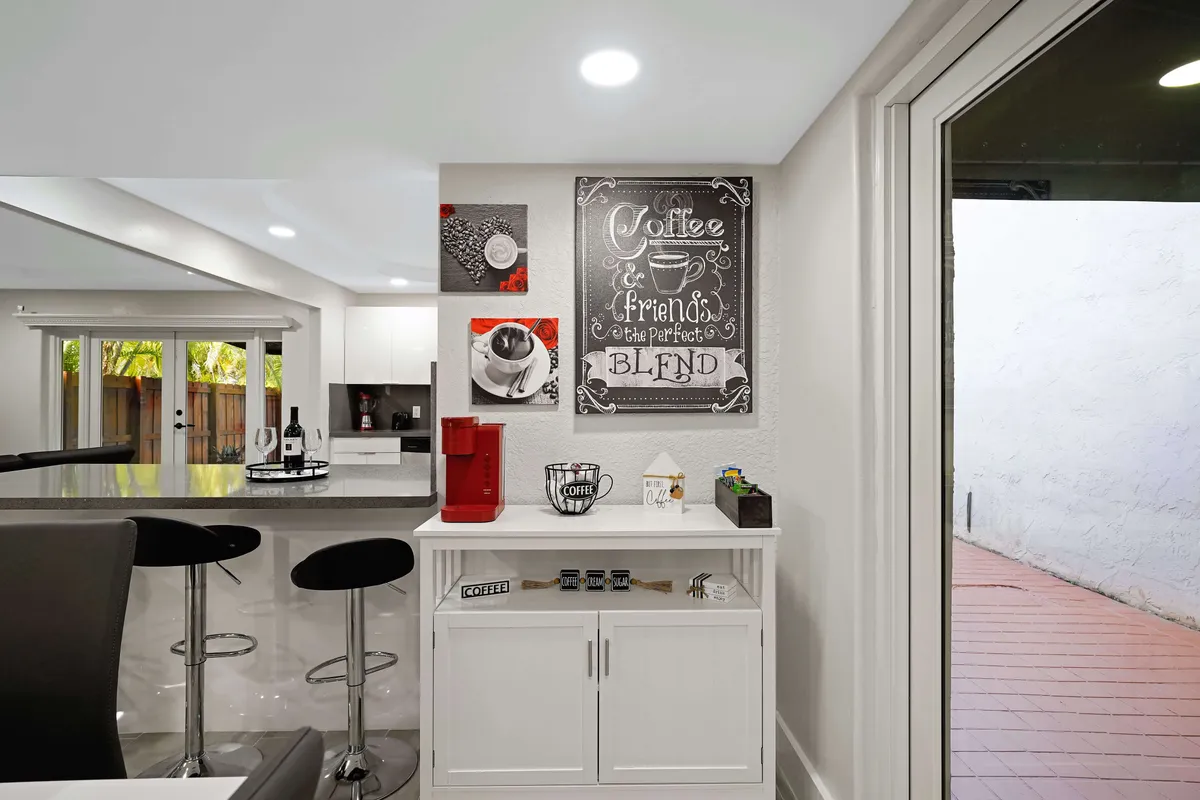 A cozy kitchen scene featuring a coffee machine, attractive artwork, bar stools, and a spacious counter. The atmosphere is warm and welcoming, ideal for enjoying coffee with friends or casual meals at the counter.