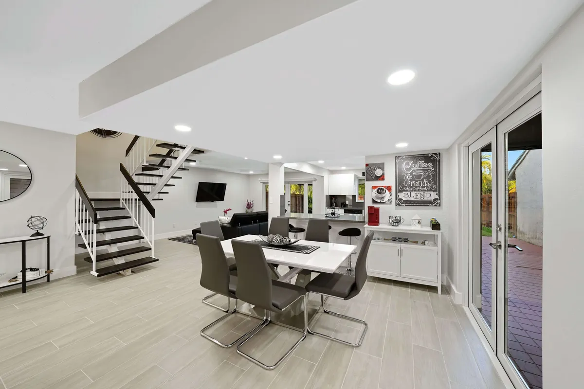 The image showcases a bright, open-concept living and dining area featuring a stylish dining table with modern chairs, a sleek staircase, a dedicated coffee station with a decorative chalkboard, and a cozy living space with a mounted smart TV. Glass doors offer a glimpse of inviting outdoor space, creating a seamless flow for entertaining or relaxing.