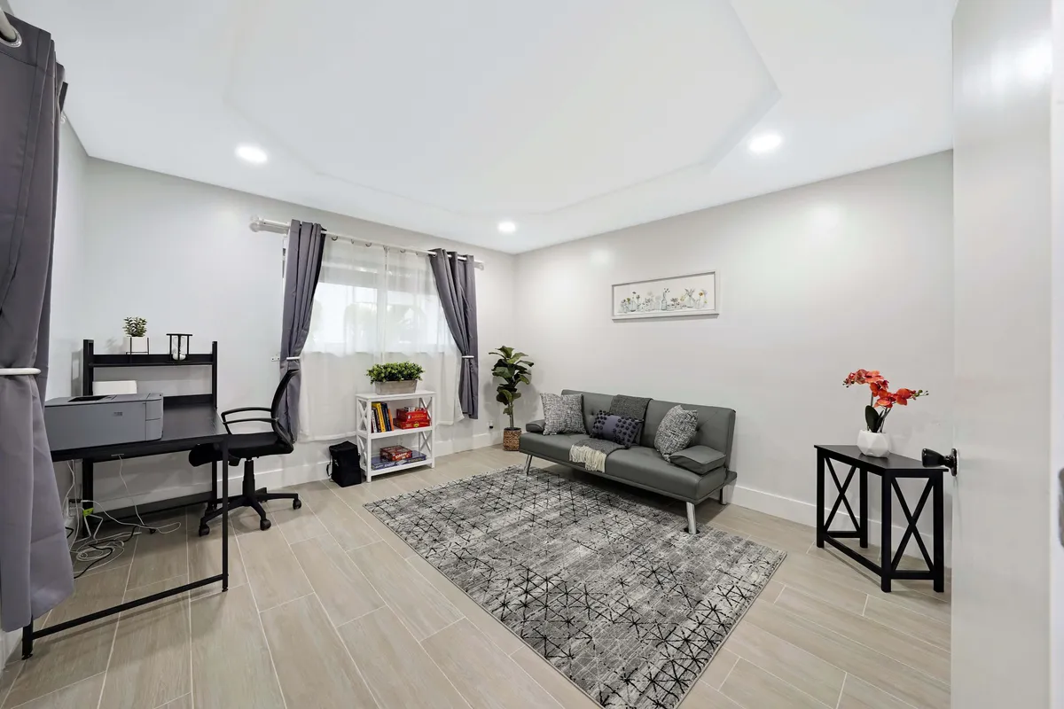 The image features a modern gray sofa with decorative pillows, a work desk with an office chair and printer, a decorative rug with geometric patterns, and a small side table topped with vibrant red flowers. The atmosphere is serene and conducive to both work and relaxation.