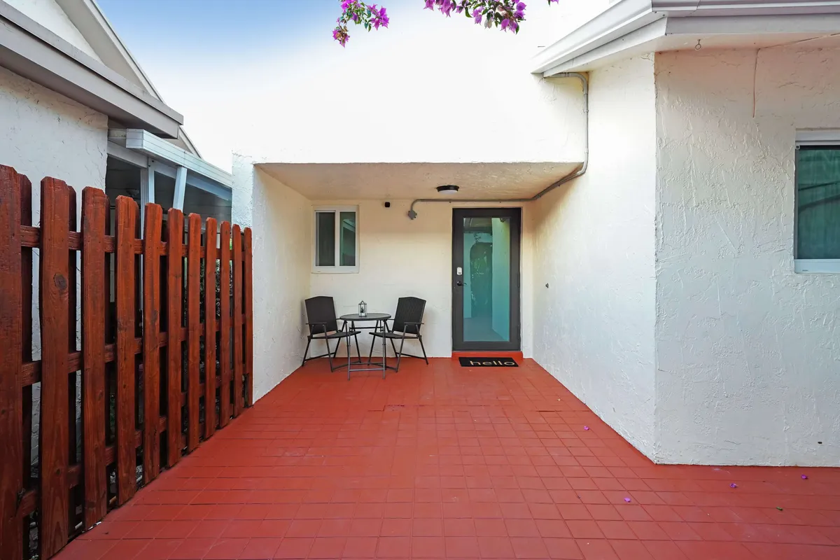 A small patio seating area with a table and two chairs sits outside a glass door entrance. A 'hello' doormat, red tiled floor, and a wooden fence provide a welcoming and private ambiance, suggesting a pleasant spot for morning coffee or conversations.