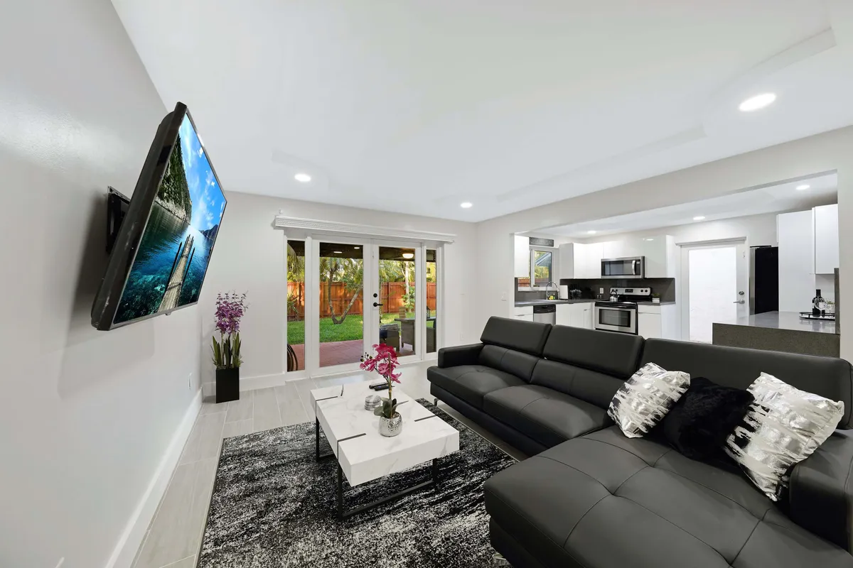 This image highlights a sleek black leather sectional sofa arranged around a modern white marble coffee table topped with orchids. The wall-mounted smart TV displays a nature scene, and large glass doors lead to a green outdoor patio, letting abundant natural light fill the space. The setting is open, stylish, and conducive to relaxation or socializing.