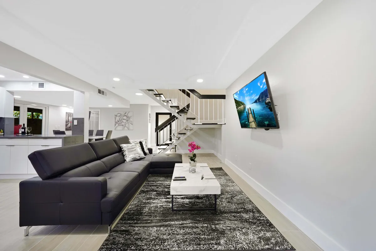 A large, black sectional sofa faces a wall-mounted smart TV displaying a scenic image. The area is accented by a modern staircase with black handrails and a white marble coffee table with pink flowers. The whole space feels contemporary, inviting, and set up for both relaxation and small gatherings.