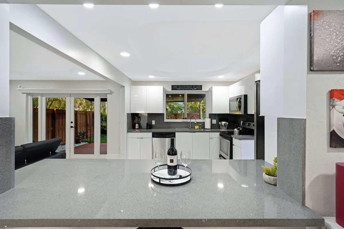 A modern kitchen with white cabinetry and appliances, a countertop set with wine and glasses, and a window above the sink that looks out onto green outdoor space. Glass sliding doors open directly to the backyard, promoting seamless indoor-outdoor living.