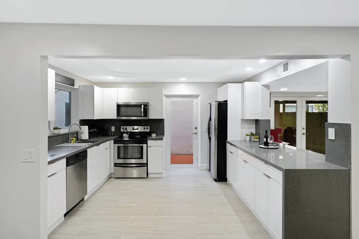 A contemporary kitchen with long quartz countertops, stainless steel appliances including oven and refrigerator, and white cabinetry. The counter overlooks an outdoor yard, and a door leads directly to the outside, making the space feel open and accessible. The setup fosters both efficient meal preparation and casual gatherings.