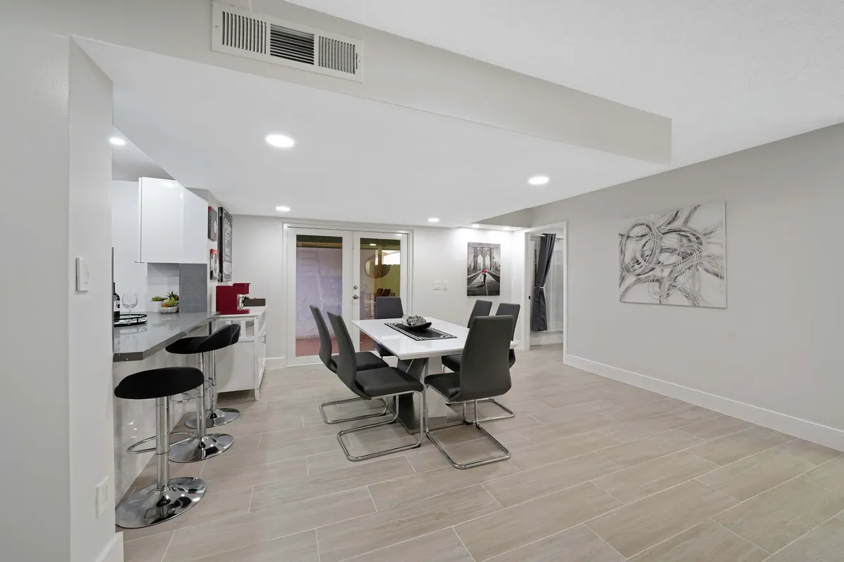 A bright, modern dining area featuring a white table surrounded by black chairs, situated next to a kitchen counter with bar stools. Abstract art decorates the wall, and glass doors provide a view outside, enhancing the space with natural light. The setup is ideal for shared meals or casual conversation.