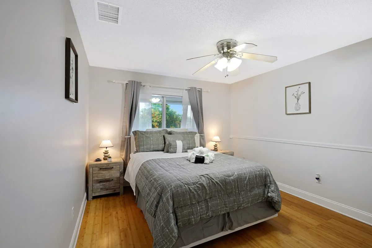 A serene master bedroom featuring a king size bed with decorative pillows and towel art, flanked by two bedside tables with lamps. A ceiling fan with lights is above, and a window with curtains overlooks lush greenery, creating a peaceful atmosphere.