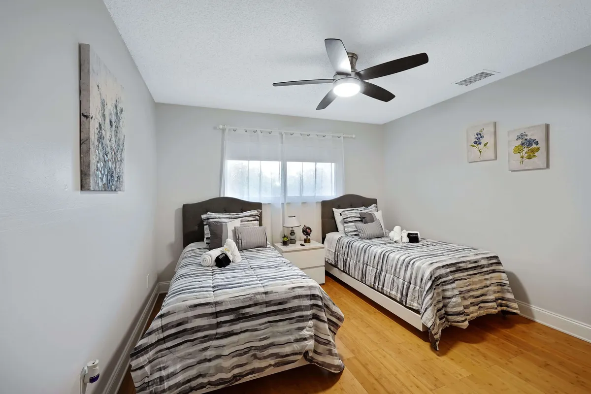 A cozy guest bedroom with two twin beds dressed in matching striped bedding. A central nightstand with a lamp sits between the beds, and floral wall art adds a gentle decorative touch. The ceiling fan and soft lighting create an inviting, restful environment.