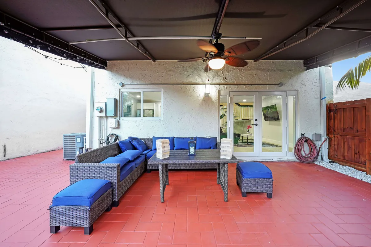 A stylish outdoor lounge setup with an L-shaped wicker sofa featuring blue cushions, a rectangular wicker table decorated for relaxation or games, a ceiling fan overhead, and large glass doors connecting to the inside. The ambiance is relaxed and sociable, ideal for casual get-togethers.