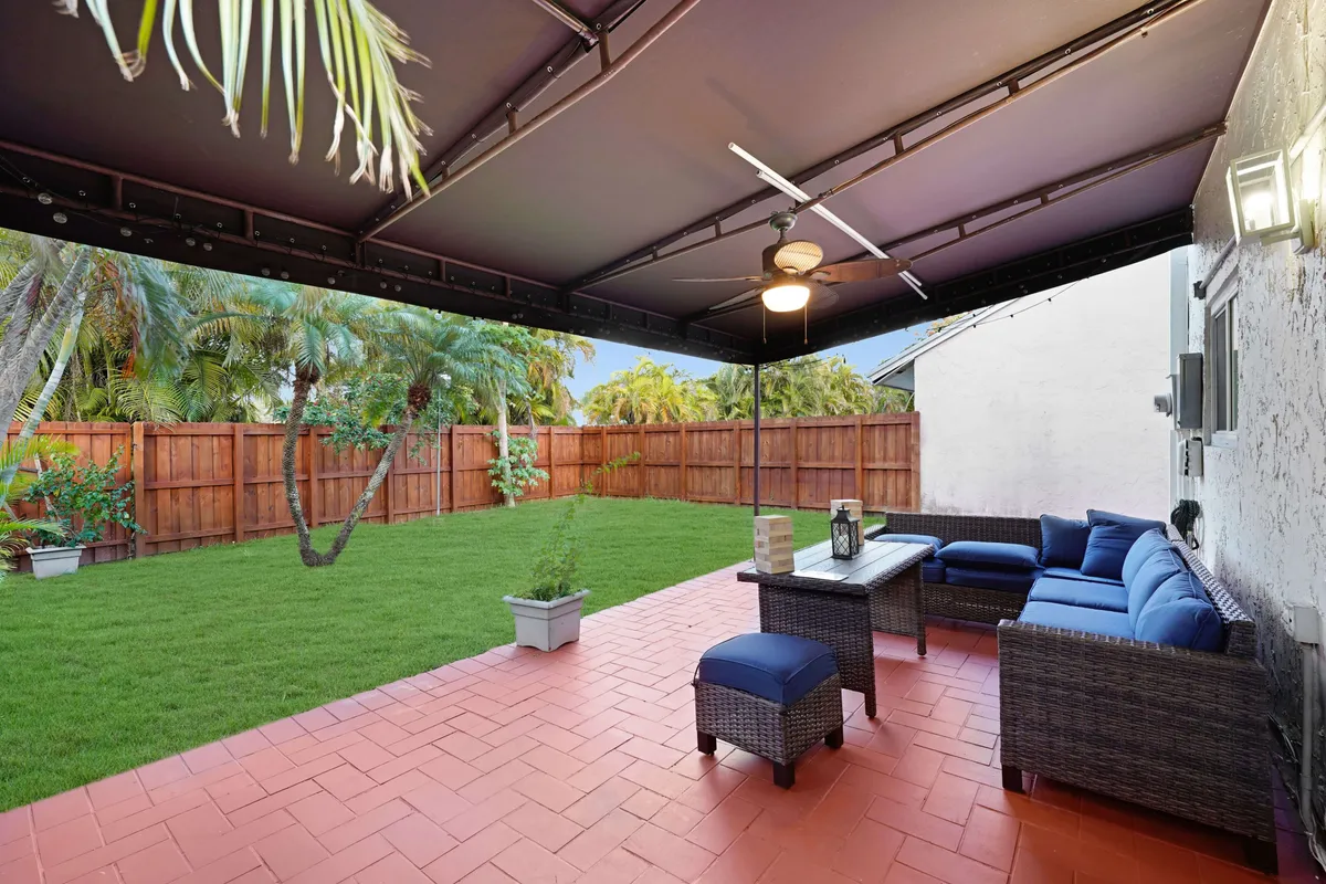 A spacious backyard with a covered patio featuring blue cushioned sectional seating, a ceiling fan, and a well-maintained lawn bordered by a wooden privacy fence. The area is ideal for relaxation, play, or outdoor gatherings.
