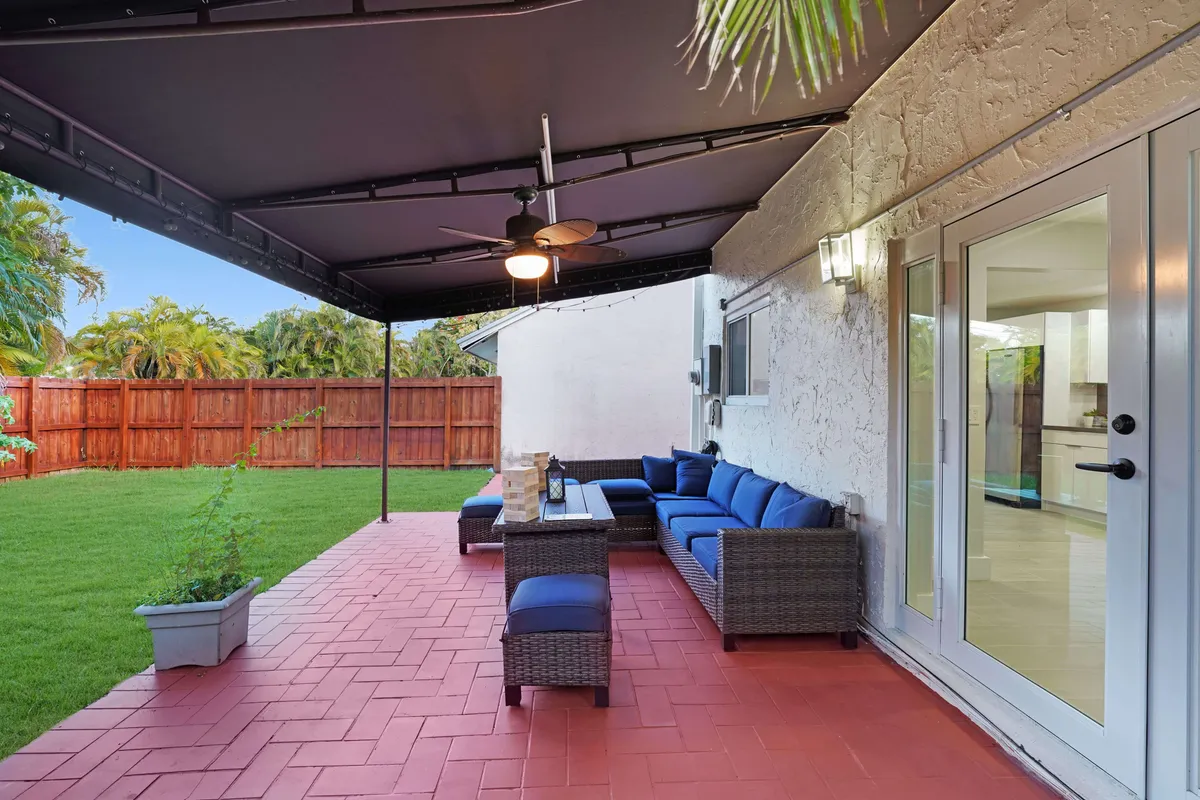 This exterior image captures a covered patio with a blue cushioned wicker sofa set, set atop red tiled flooring and surrounded by a well-kept green lawn and wooden fence. Glass double doors offer a seamless transition between interior workspaces and the tranquil outdoor environment.