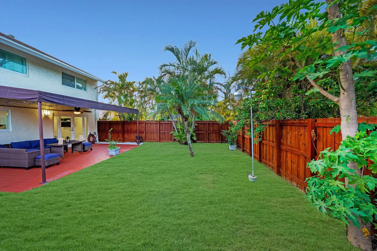 A wide view of a vibrant green lawn surrounded by tropical plants and palm trees. The covered patio seating area provides shade and comfort, making it an appealing spot for family relaxation, BBQs, or group activities. The wooden privacy fence ensures a safe space for children.