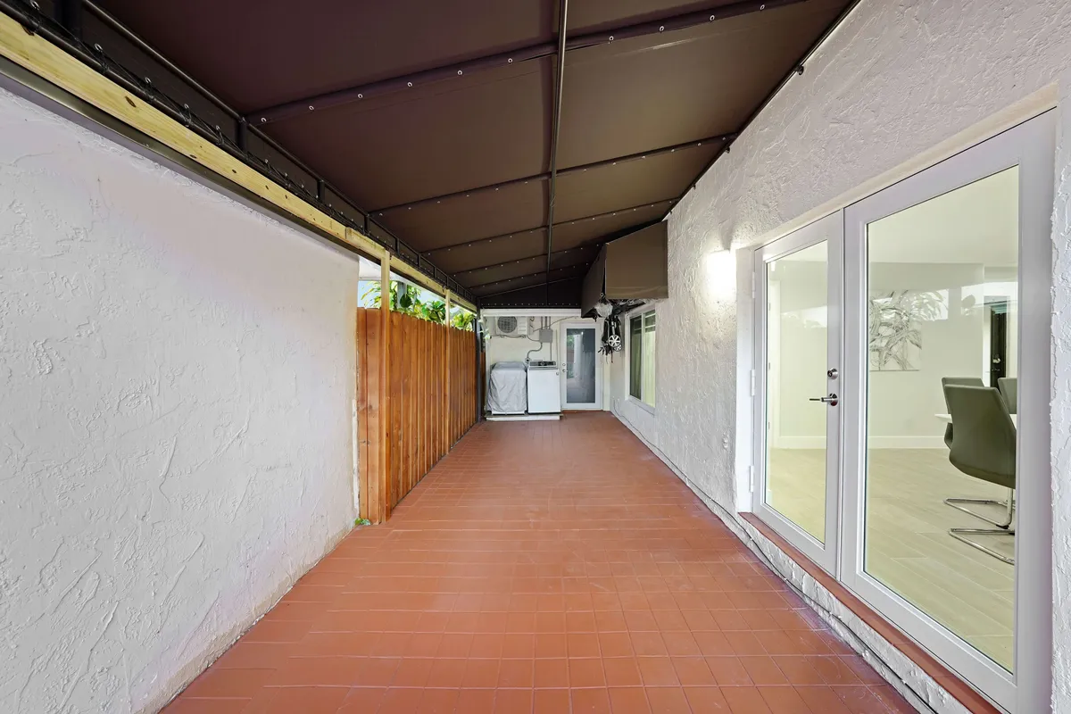 A covered patio area with terracotta tile flooring leads to a laundry area featuring a full-size washer and dryer. The space is bordered by a wooden privacy fence and connects to the indoor living area through glass doors.