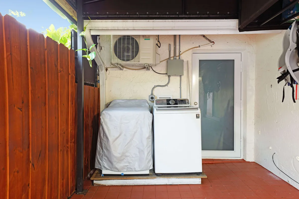 A practical laundry area featuring a washing machine with a protective cover and a dryer unit, set next to a glass door that opens to the outdoors. An air conditioning unit is mounted on the wall, hinting at comfort and year-round utility. The setup is both convenient and accessible, ideal for longer stays.