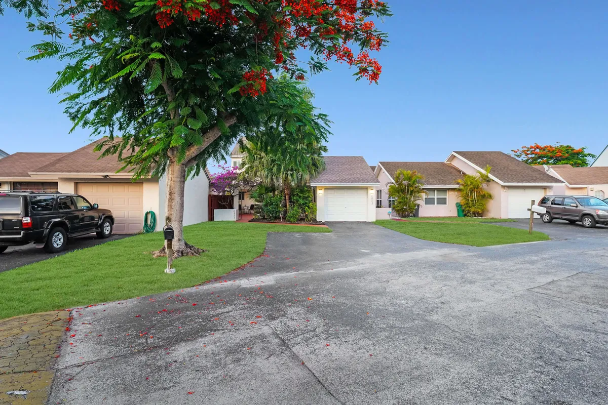 A tranquil neighborhood scene showing a row of single-story homes with garages, lush green lawns, a large tree with red blossoms, and parked cars along the driveway. The image evokes a sense of arrival and the peaceful, welcoming character of the surrounding area.