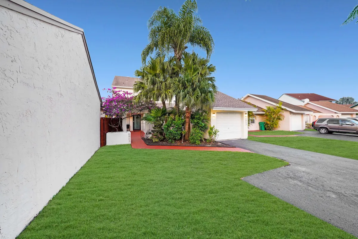 A view of the well-maintained front lawn, framed by tall palm trees and lush greenery, leads up to the house entrance via a red pathway. An attached garage and the quiet residential street hint at convenient access for travelers and a tranquil return after exploring nearby attractions.