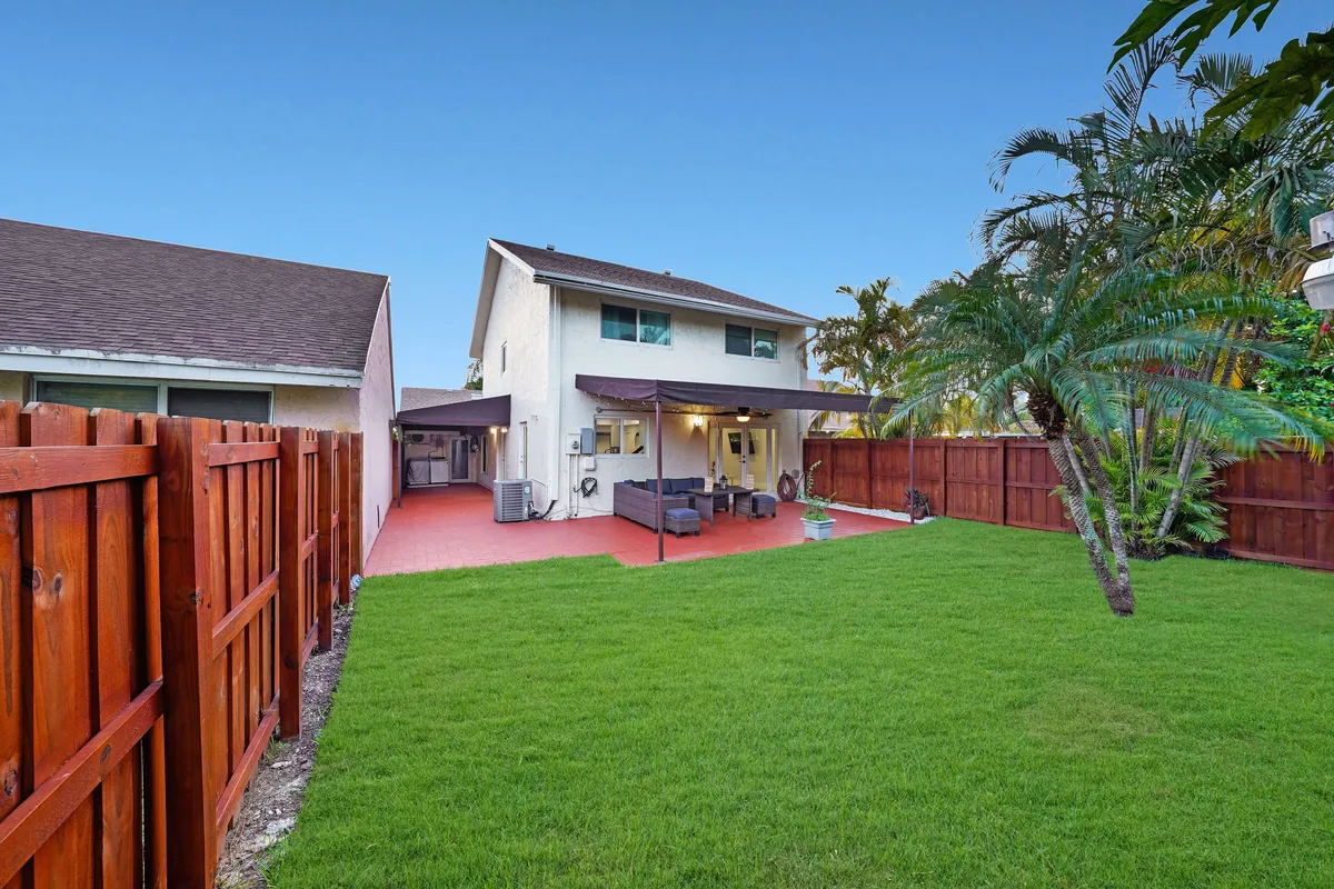 A spacious backyard featuring a large green lawn bordered by a wooden fence, with tall palm trees adding a tropical touch. The covered patio is furnished with outdoor seating, inviting for gatherings or relaxation. The area is sunlit and ideal for family activities, outdoor games, or barbecues.