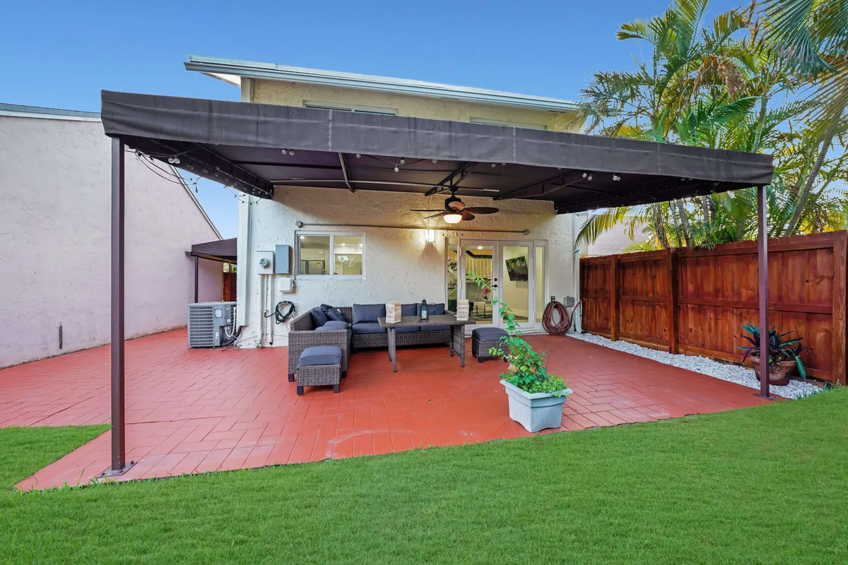 A welcoming covered patio area featuring a sectional sofa with cushions, a ceiling fan, string lights, and potted plants. The patio floor is tiled in red, contrasting with the adjacent green lawn. The setting seamlessly blends indoor comfort with outdoor leisure, creating an inviting social space.
